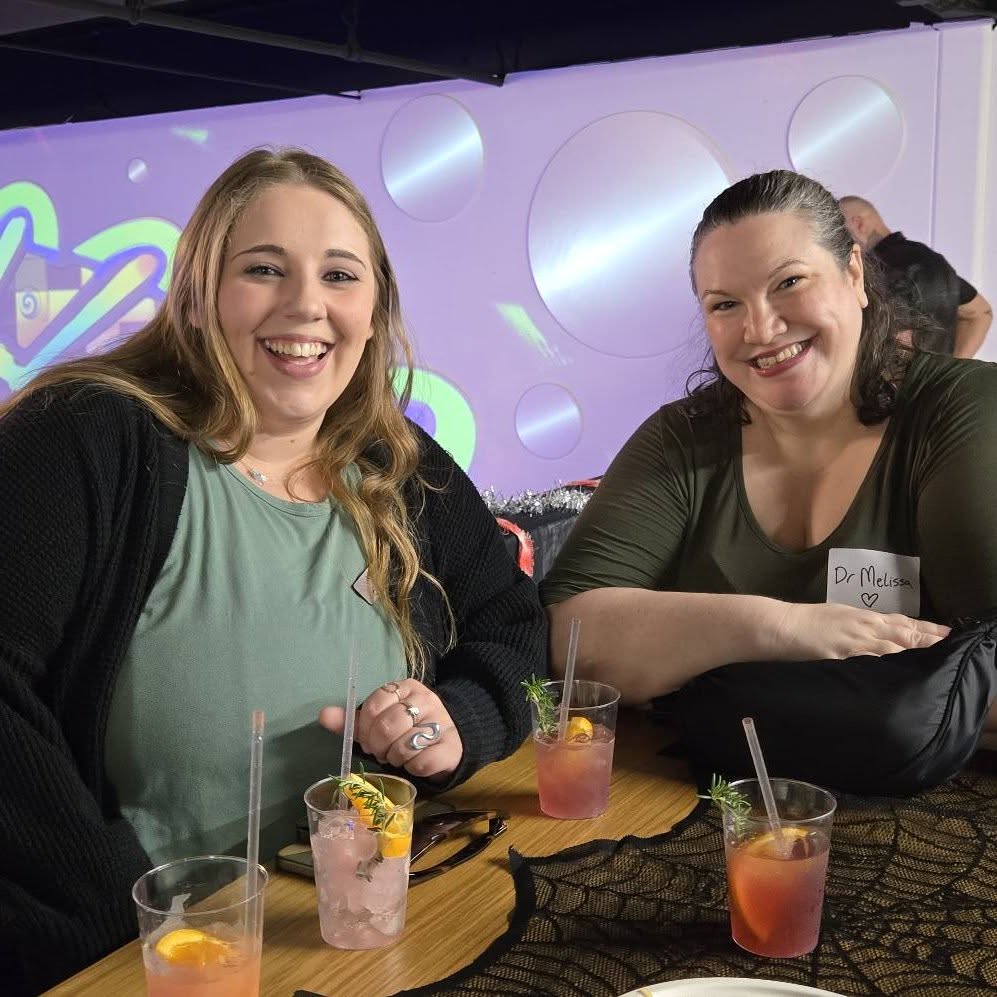 Two women smile while sitting at a table with drinks at a networking mixer.